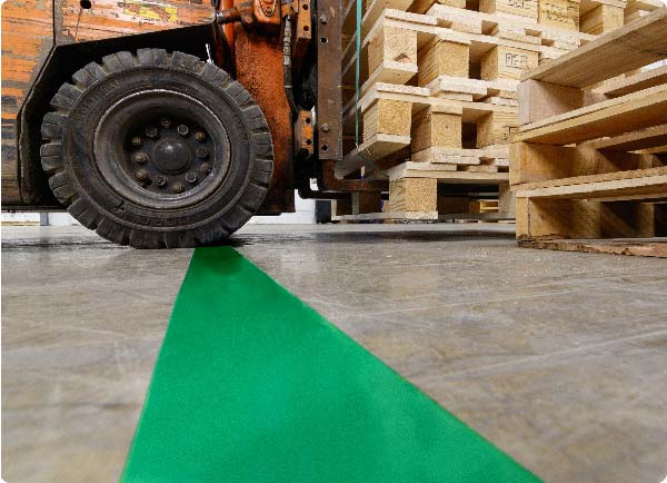 Een grote vorkheftruck rijdt over een lijn groene ToughStripe Max-vloertape. De randen van de tape worden niet beschadigd door de zware wielen van de vorkheftruck.