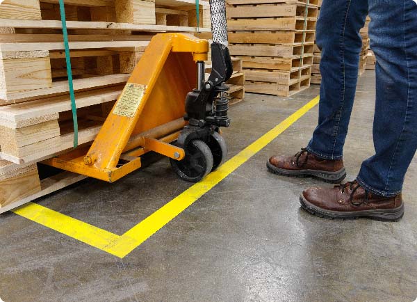 Felgele ToughStripe-vloertape wordt aangebracht op een magazijnvloer. De positie geeft aan waar de operator van een palletstapelaar een stapel materialen moet neerleggen.