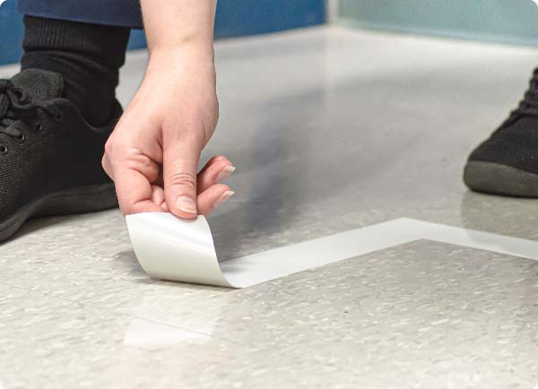 Een persoon gebruikt zijn handen om ToughStripe Vinyl-vloertape te verwijderen van vinyltegels. De tape laat zich in één stuk verwijderen.