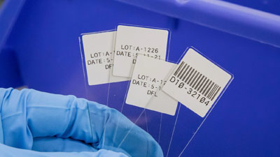 Een laborant houdt verschillende objectglaasjes vast met witte StainerBondz-labels, sommige met tekst en één ook met een geprinte barcode.
